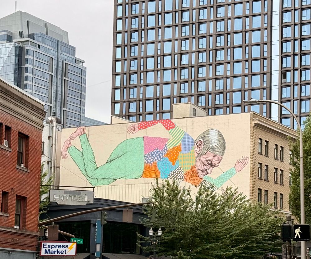 Mural of an older person in a multi colored shirt and green pants on the side of a building in downtown Portland.