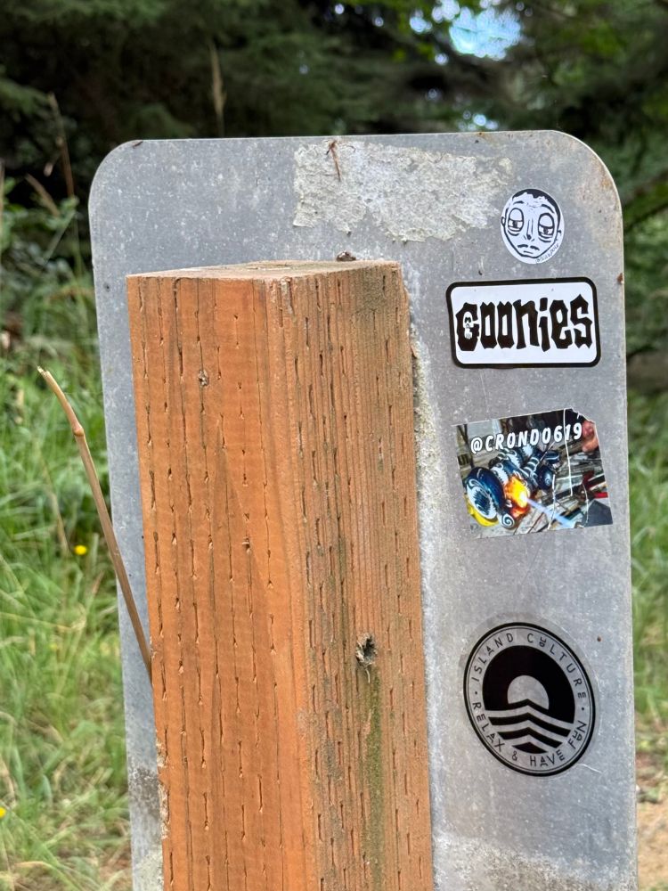 Back of a road sign with stickers on it. One of them is GOONIES.