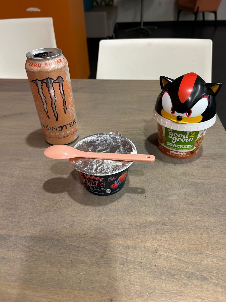A peach flavored Monster can, graham crackers with a Shadow the Hedgehog lid, and a mixed berry protein yogurt sit on a faux wood table. There’s a pink plastic spoon sitting on top of the yogurt.