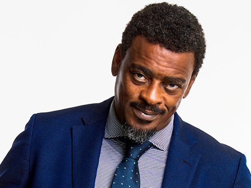 A foto mostra o cantor Seu Jorge (homem preto, de bigode e cavanhaque um pouco grisalho, vestindo um paletó azul sobre uma camisa social listrada, com gravata também na cor azul) olhando para a câmera com uma expressão facial sorrateira.