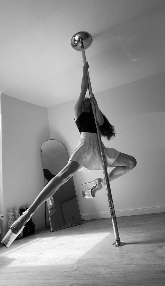 A black and white screenshot of Val frozen in mid air as she pole dances. Her right leg is extended and her left leg is bent as she hangs from the pole. She is wearing a white pleated skirt and black tank top. Her hair is long and brunette. 
