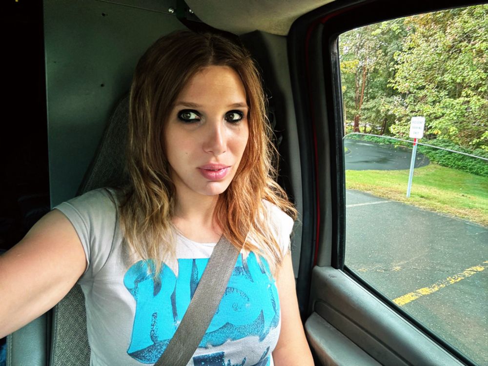 Blond woman sitting in van looking youthful wearing a rush shirt. Late 20s