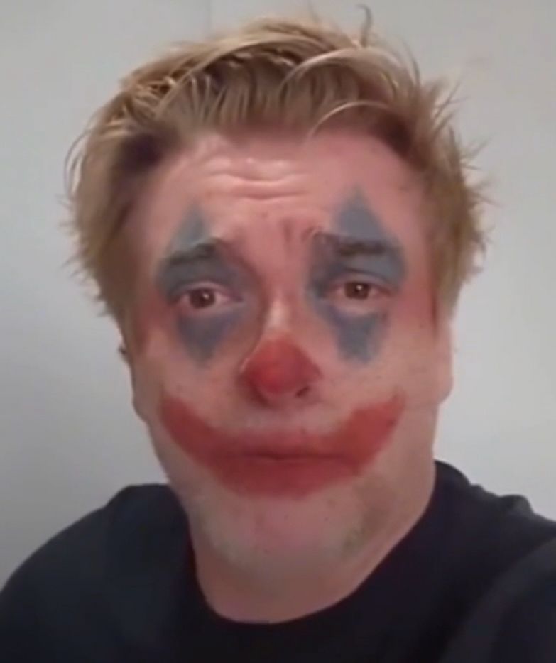 A screenshot of Rhys Darby from one of his videos, but a clown makeup filter has been put on his face. He’s subsequently looking into the camera with a red nose, an overpainted red mouth, and blue triangles over his eyes.