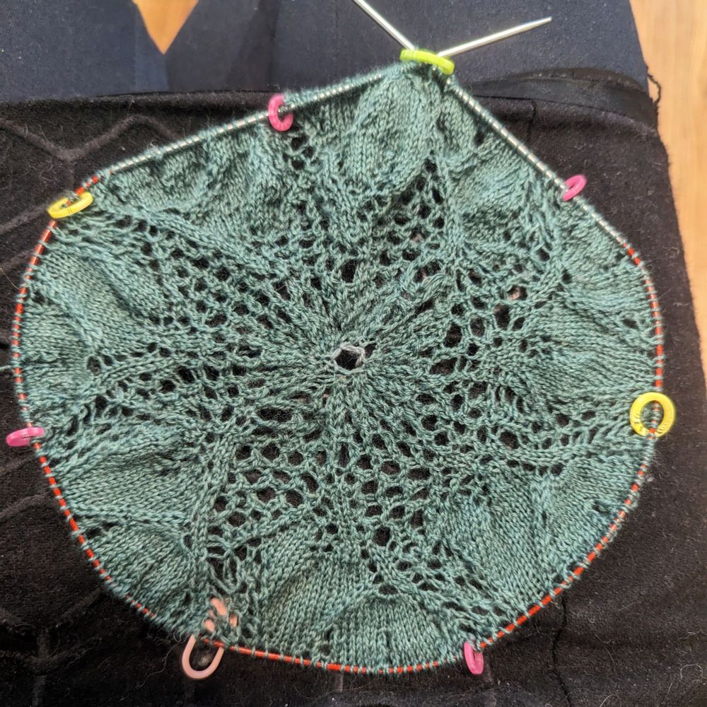 A messy half-finished Niebling lace doily sitting in the lap of the knitter. It's knitted in a blue-green wool.