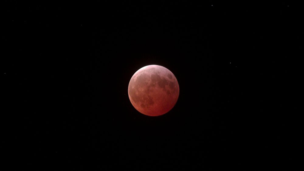 The full moon during totality during the lunar eclipse on March 13-14, 2025. The moon is colored deep red with a bit of lightness at the top. There are a few stars visible in the background.
