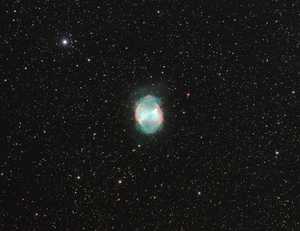 A oval nebula with the longer dimension vertical is in the center of the image. It is mostly blue-green with reddish colors on the left and right and a bit at the top and mixed in tangles through the nebula. The nebula is set against a dense field of thousands of stars, with one brighter star to the upper left.