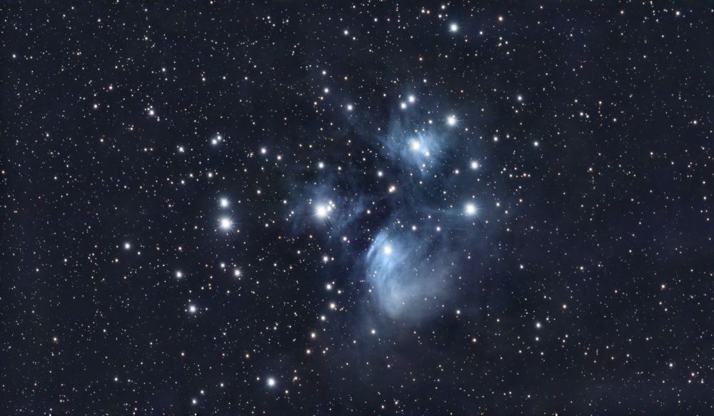 A cluster of bright stars filtered through clouds of bluish white dust in the center of the image, set against a background field of dimmer stars.