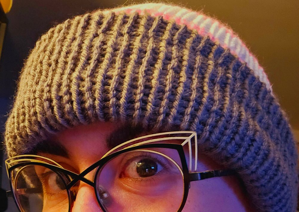 Photo of the top half of a white woman's face with dark brows wearing cat eye glasses and a demigirl striped knitted beanie hat.
