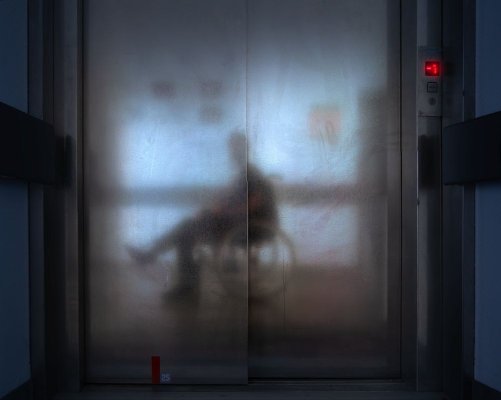 Eng : Color photo of the reflection of a man in a wheelchair in the metal door of an elevator.

Fr : Photo en couleur du reflet d'un homme en fauteuil roulant dans la porte métallique d'un ascenseur.