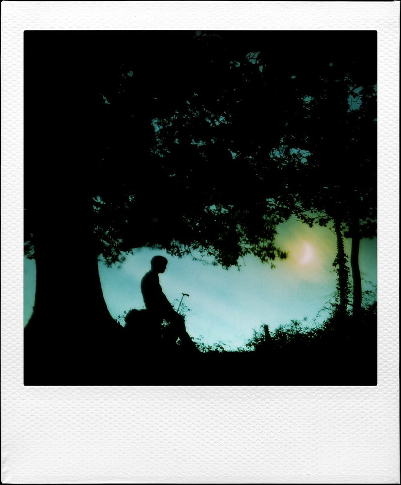 Photo polaroid d'un paysage nocturne.
La silhouette d'un homme se tient sous un arbre, il est assis sur une souche et tient une canne de marche dans sa main.
Dans le ciel nuageux et bleuté, un croissant de Lune est visible dans un large halo jaune.