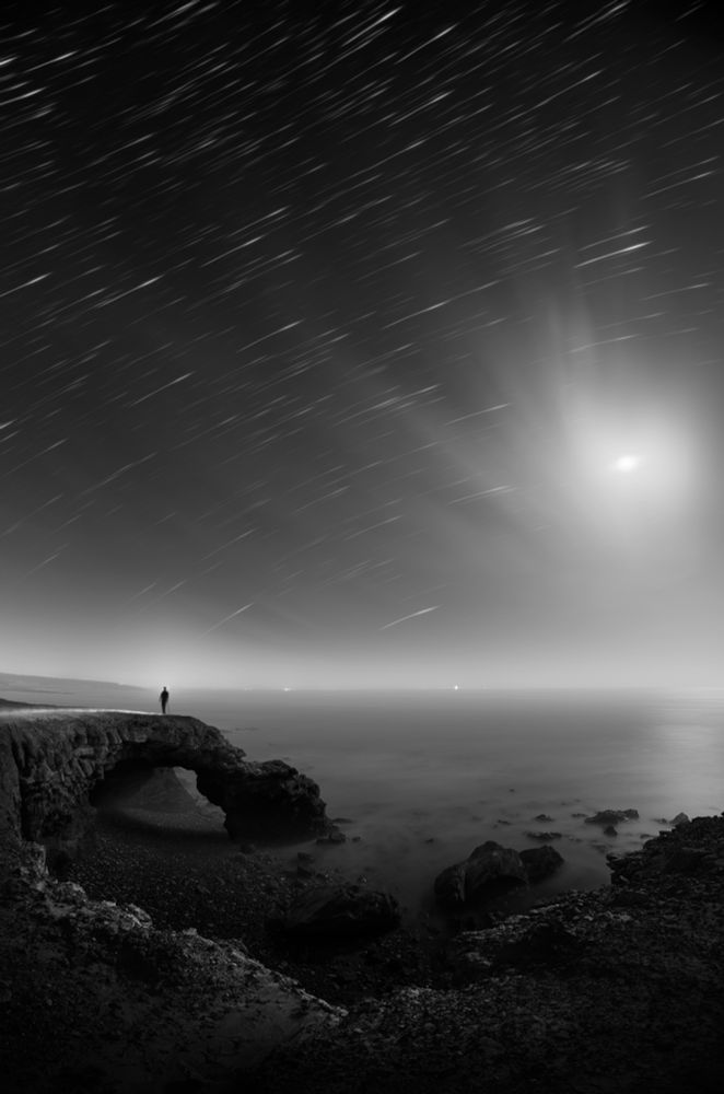 Eng ; Photo of a night landscape in black and white.
At the edge of a cliff, a man observes the sea under the stars, standing on a rock bridge.
Above him, the long exposure reveals the stars as lights that seem to drift away.

Fr : Photo d'un paysage nocturne en noir et blanc.
Au bord d'une falaise, un homme observe la mer, sous des étoiles, perché sur un pont de roche.
Au dessus de lui la pose longue fait apparaitre les étoiles commes de lumieres qui semblent filer.