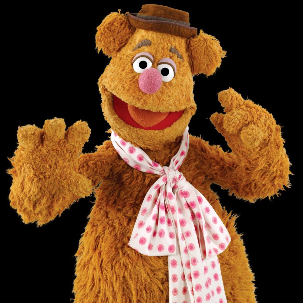 Brown-furred puppet with his mouth open wearing a polka-dotted scarf and brown porkpie hat