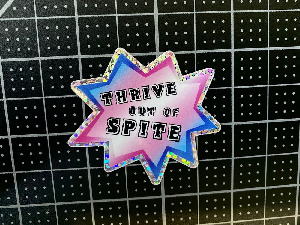 Photograph of a sticker against a black and white background. The sticker features the text "Thrive out of Spite" against a trans flag gradient background.