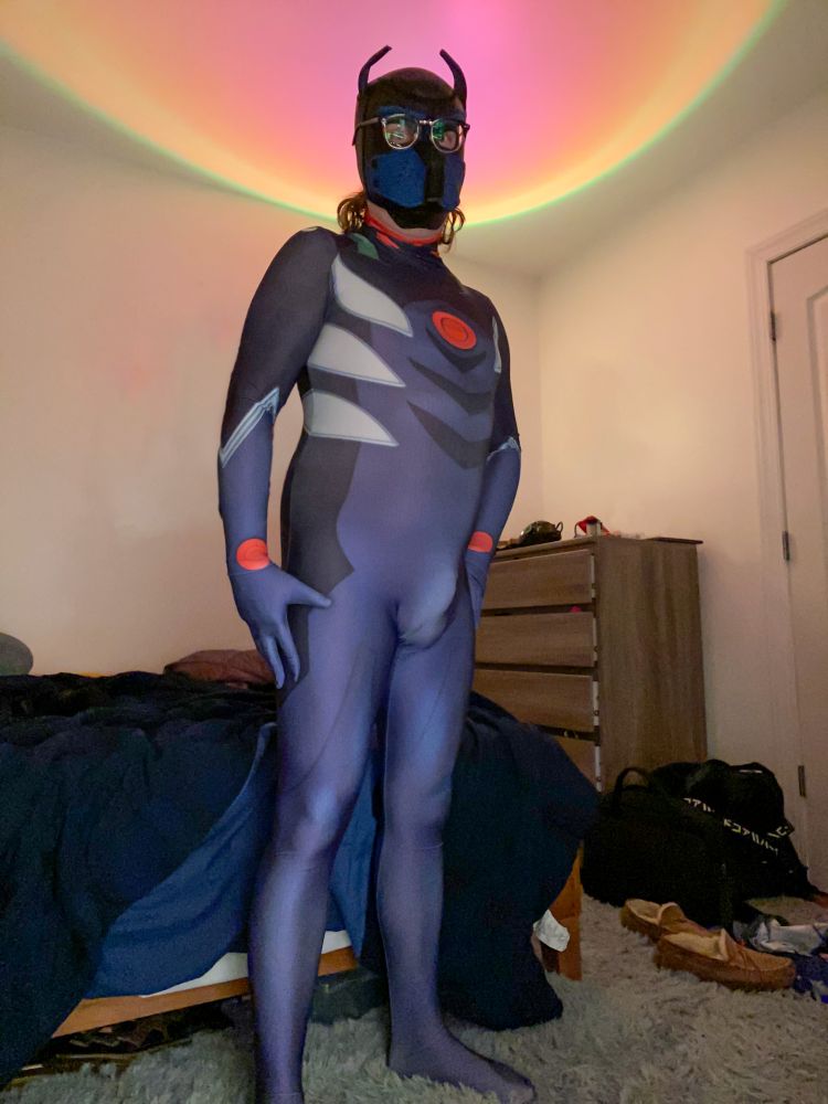 Picture of a person wearing standing in a slightly cluttered room. They are wearing a black and blue pup hood, glasses, and a spandex suit reminiscent of Kaworu's plugsuit from Evangelion.