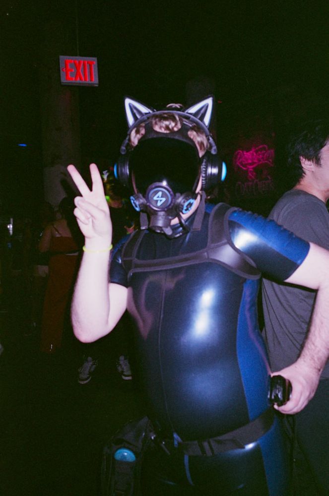 Film photograph of a drone wearing a metallic dark grey and metallic blue surfsuit with a neoprene harness. They are holding up a peace sign with one hand.