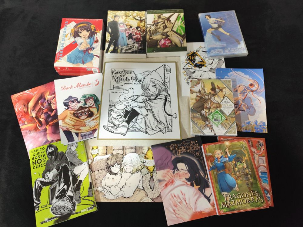 Fotografia sobre fons negre on es veuen diversos còmics, caixes de DVD i cartulines artístiques. Al centre de la imatge hi ha un dibuix de Coco, la bruixa protagonista del manga 'Atelier of Witch Hat', signada per l'autora, Kamome Shirahama. A la part superior es veuen, d'esquerra a dreta, els DVD de 'Haruhi Suzumiya', 'Steins;Gate 0', 'La visión de Escaflowne' i 'La chica que salta a través del tiempo'. A l'esquerra, una print de Pokémon i el fanzine 'Dark Limbo 3'. A la dreta, el volum 13 del manga 'Atelier of Witch Hat', una print de la mateixa sèrie i una print de 'Frieren'. A la part de baix, el manga 'El chico que me gusta no es un chico', una altra print de 'Atelier of Witch Hat', el fanzine 'Renée' i els volums 2 i 3 del manga 'Tragones y Mazmorras'.