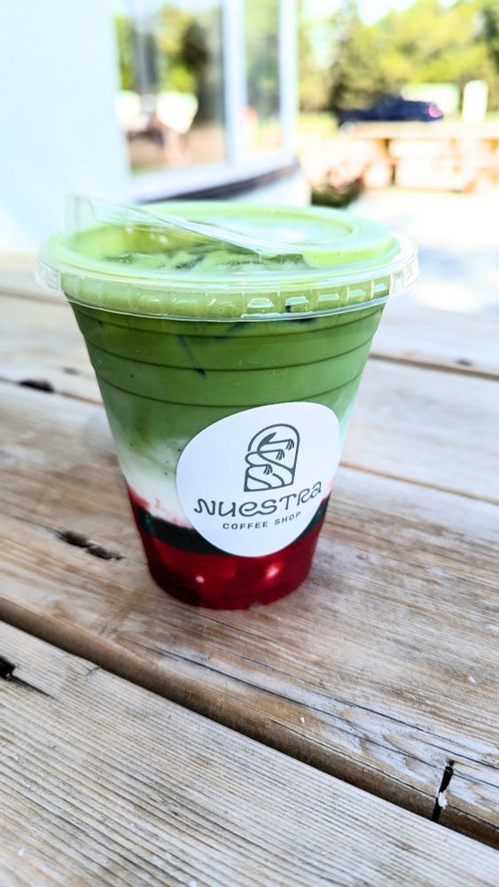 A clear plastic cup on a picnic table in front of a cafe. In the cup is a mixed berry matcha, with a bright red berry sauce on the bottom. There's a round white sticker on the cup with the name of the coffee shop (Nuestra).