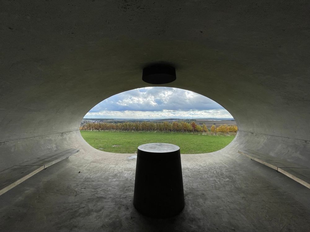 Blick aus einer nierenförmigen Betonröhre (die flache Seite ist unten) auf eine Landschaft mit Weinbergen. An der Wand geht eine Holzbank entlang, direkt vor der Öffnung steht ein runder Zylinder, der auch als kleiner Tisch dienen kann.