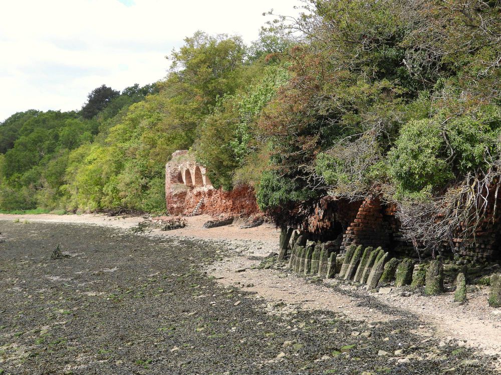 Among greenery, the red brickwork of the crumbling Cockham Wood Fort (1669). Foreground, right, a line of old defensive stakes shows high tides reach the fort (covering the England Coast Path here, diverting walkers inland). The neap tide height is well illustrated by seaweed growth on the beach.