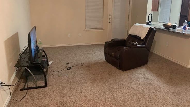 Sparsely furnished living room consisting solely of an entertainment center with exposed wires, a leather recliner with a white pillowcase for a headrest, and a discarded gaming controller stretched between the two.