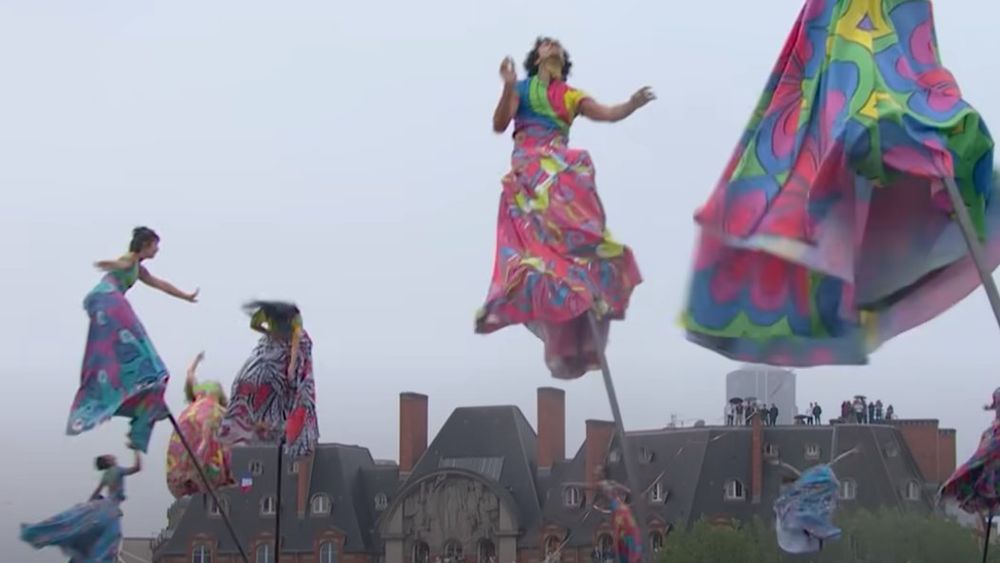 Acrobatic ballet dancers are swinging back and forth in the air atop flexible poles while their colorful flowing robes trail behind them