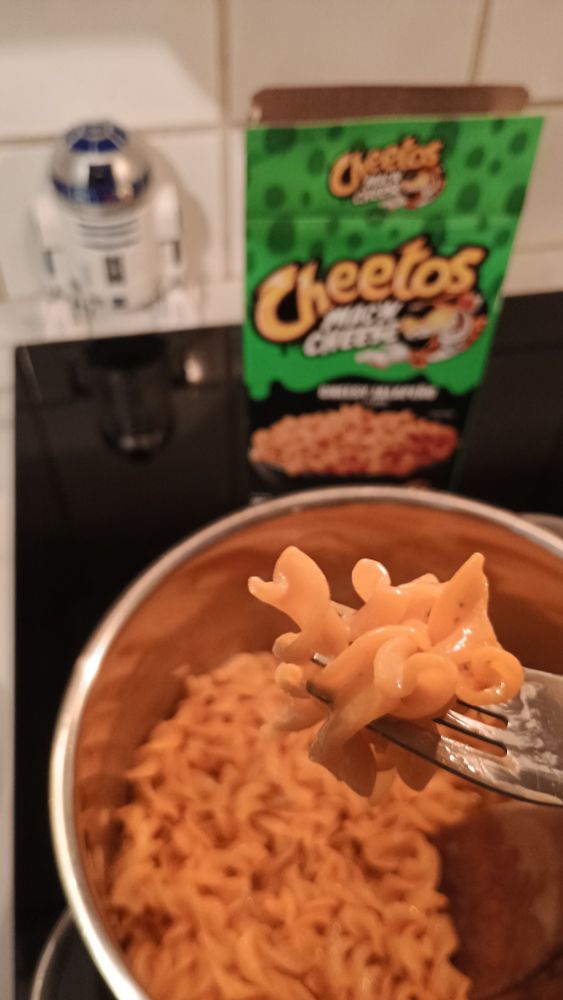 Photo of a pot of a freshly-cooked pot of Cheetos brand mac & cheese. The box is visible in the background, and a fork lifts a few noodles closer to the camera. The cheesy pasta has Cheetos' unnaturally vibrant orange color. At the back of the stove, an R2-D2 figurine looks on disapprovingly.