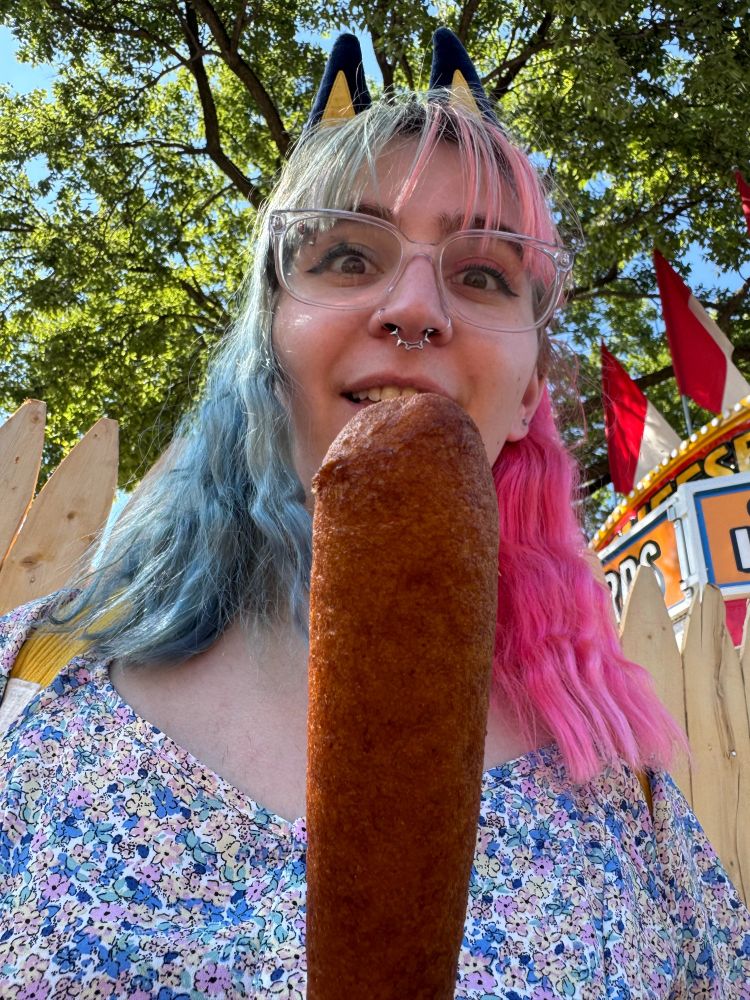 close up picture of juni from head to shoulders making big eyes at a pronto pup (looks like a corn dog) right in front of her mouth 