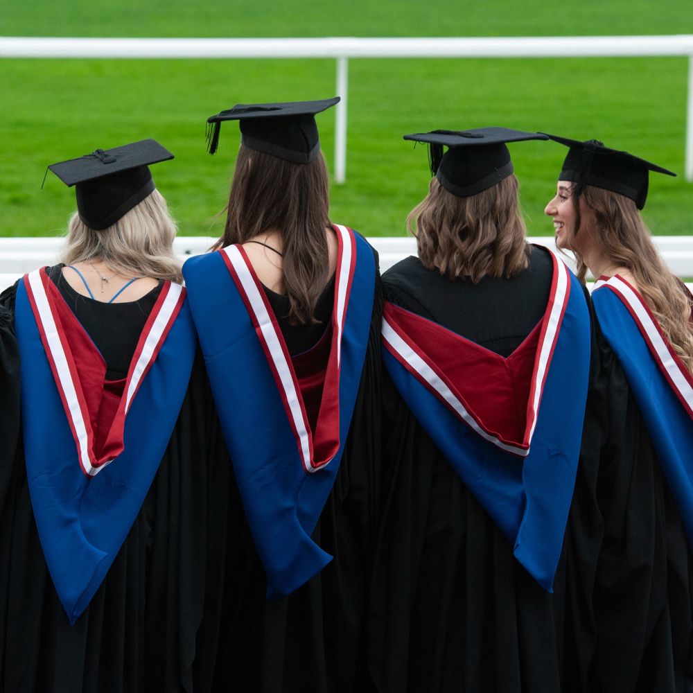 4 graduates in their gowns facing out to the racecourse