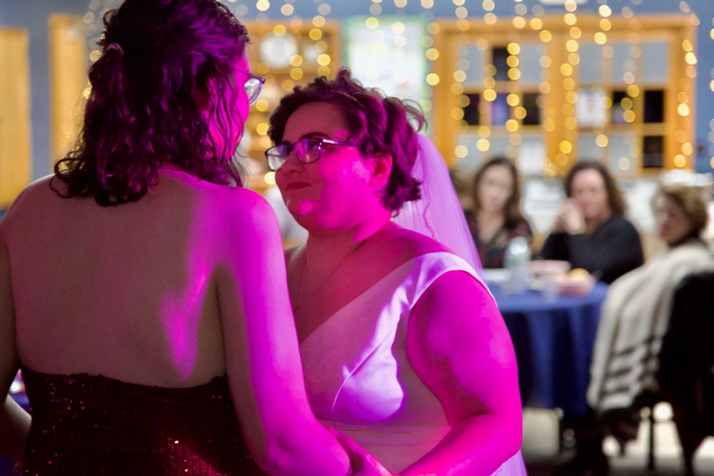 The married couple dancing with each other with fairy lights in the background as guests (including a lesbian couple) watch on