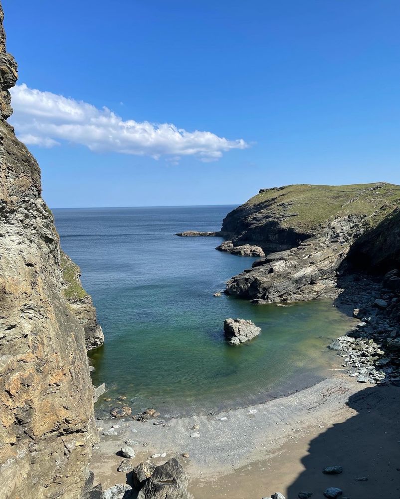 A photograph of a coastal inlet with outcroppings of rock and a small beach on a sunny day. The sky is blue and clear except for one long, fluffy white cloud.