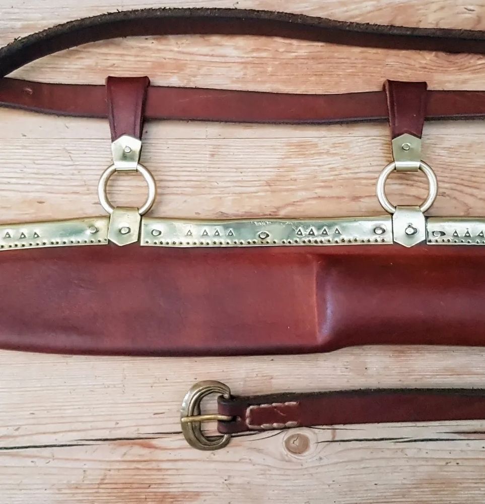 Photo of a seax sheath, made of leather and worked brass, with an attached belt. It's laying on a wooden surface. 