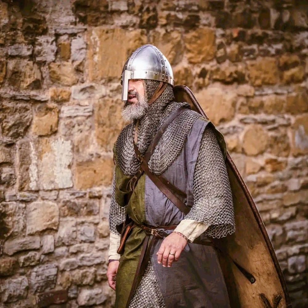 Photo of a bearded man dressed in medieval martial clothing, mail and helmet, walking past a stone wall. 