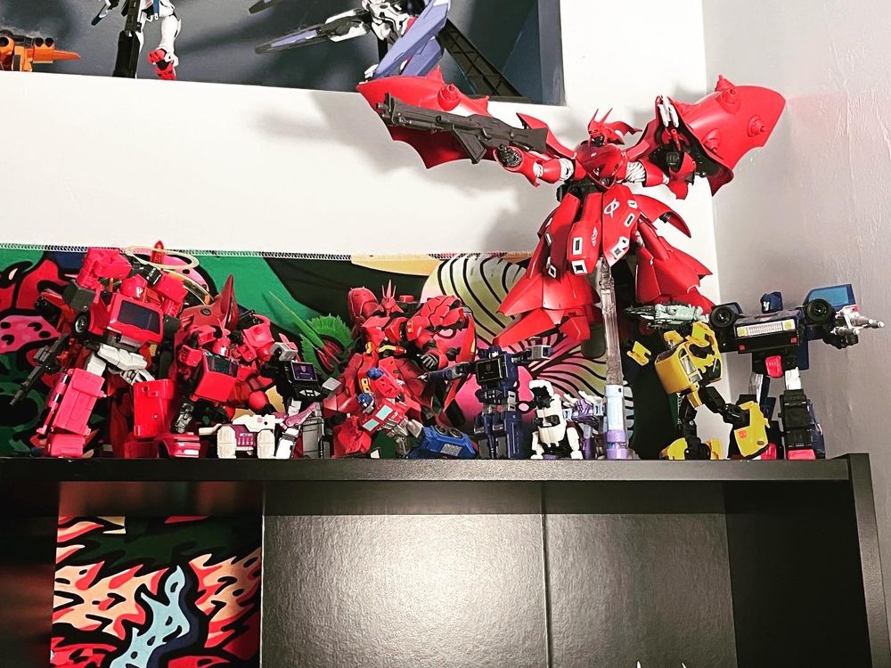Assorted colors of red robots line the top of a black shelf. The background is white, several more robots are perched on a higher shelf behind.