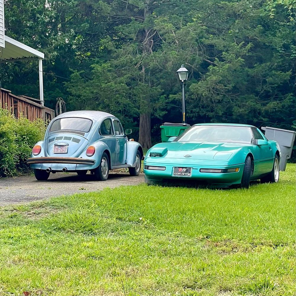 Old 70s(?) blue VW beetle next to an 80s era Corvette in teal. One of the Corvettes pop up headlights is slightly up.