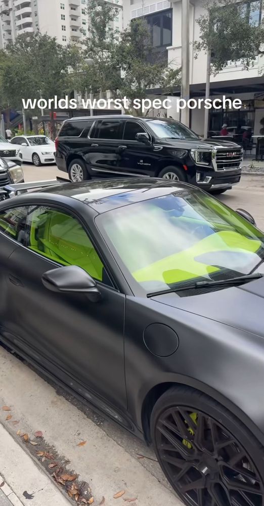 A satin black 911 with a bright yellow interior parked on street. Caption from OP says “worlds worst spec Porsche”