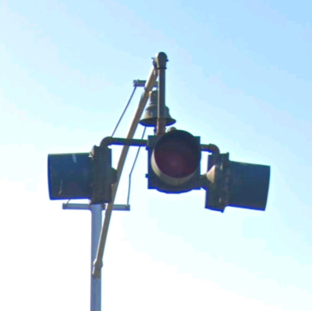 Three single traffic light heads on an old thin post with a guy wire. There is a bell hanging above and between them.