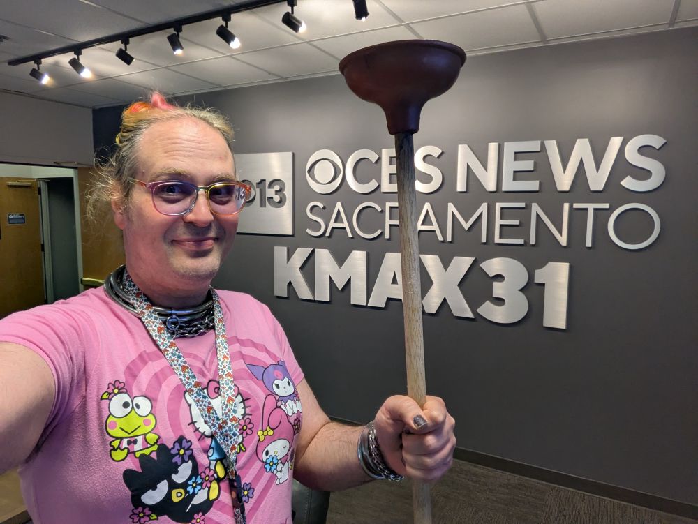 A person standing in front of a TV station's logos on the wall, holding a plunger. They're wearing a cute Hello Kitty and Friends shirt.