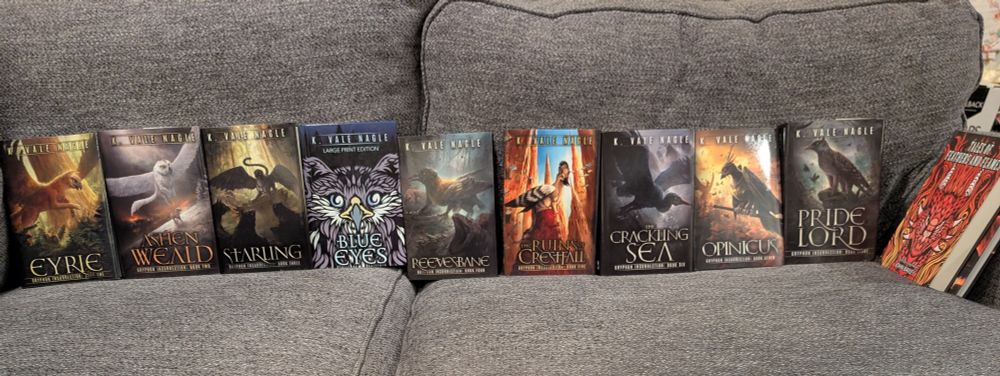 A photo cropped wide of a couch with ten hardcover 6" x 9" epic fantasy books in a row, face out. They all have gryphons on them except the last, which has a wyvern. The books, as displayed, are Eyrie, Ashen Weald, Starling, Blue Eyes and Other Tales, Reevesbane, The Ruins of Crestfall, The Crackling Sea, Opinicus, Pridelord, and Tales of Feathers and Flames. Behind Feathers and Flames is a partially visible hardcover of Dire by John Bailey.