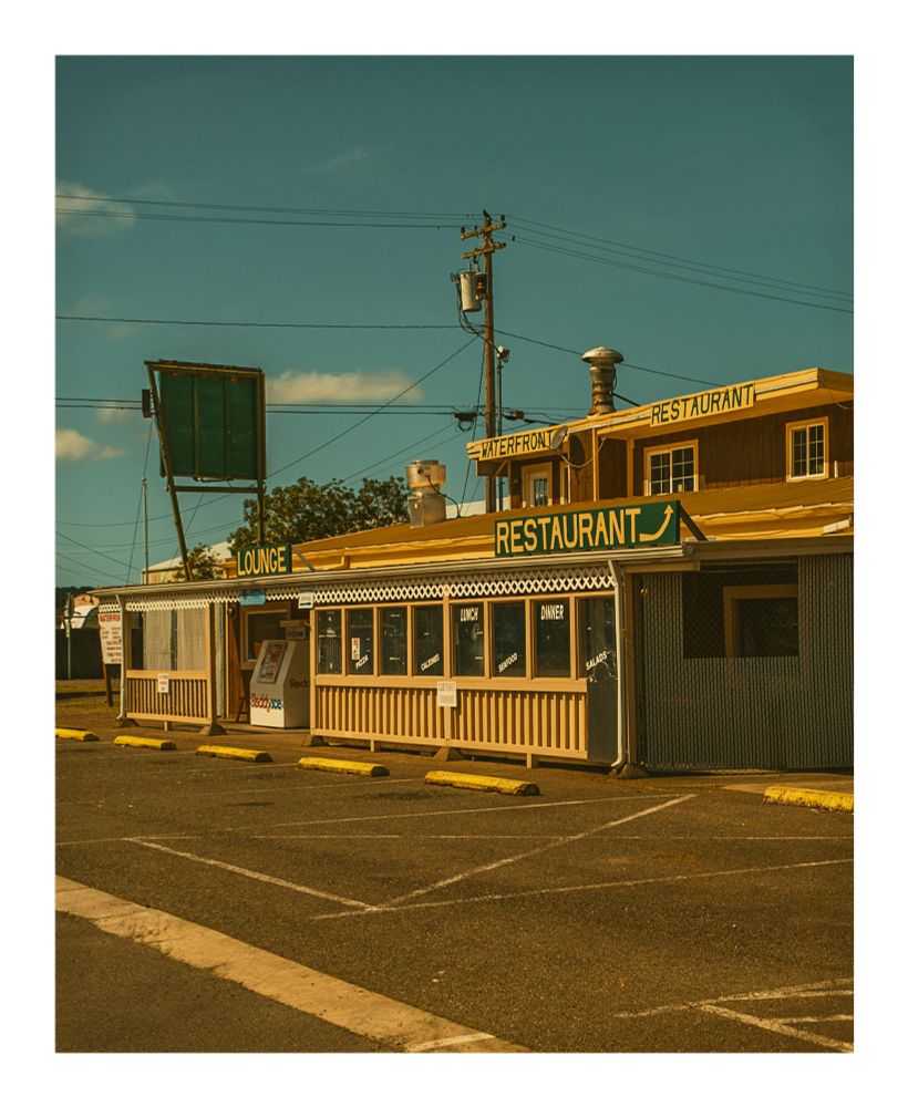 A color photograph of Kismet’s Landing restaurant in Reedsport, Oregon. It is a yellow painted building, with hand painted restaurant signage. The Peking lot is empty.