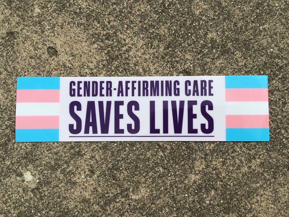 A bumper sticker that says “gender-affirming care saves lives” in dark purple text on an off-white, purple tinted background. On either side of the text is the trans flag, which has light blue, light pink, and white stripes.