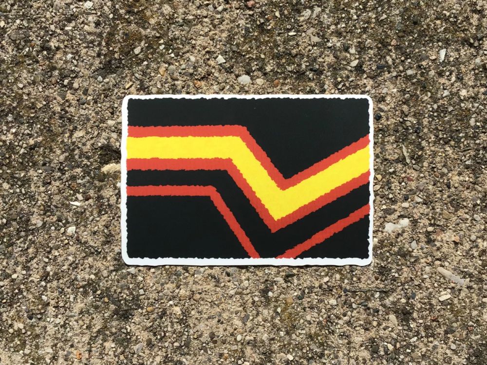 A photo of a sticker. The sticker has a thin white border. The design is of the Rubber Kink pride flag: a black flag with a series of crooked yellow and red stripes.
