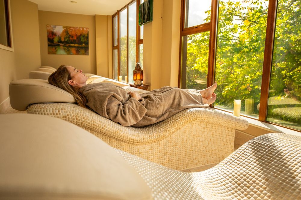 A woman with long blonde hair wearing a beige towelling robe lies on a couch in Ireland's Falls Hotel & Spa's relaxation room, facing green woodland seen through high indows, and bathed in soft golden sunlight. 