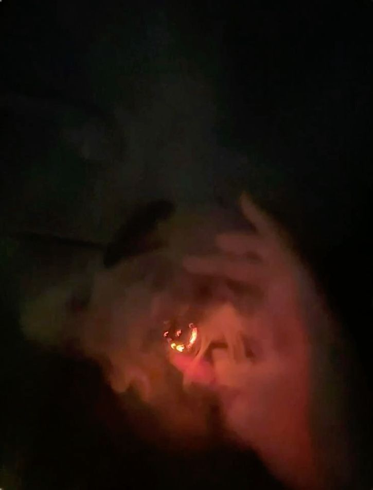 A dark, hazy image of someone holding a lit blunt the ember accentuating the smoke, red lighting, and barely visible hand 