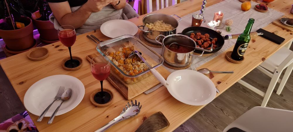 Ein für drei Personen gedeckten Tisch mit Töpfen, einer Pfanne, Kerzen und hinten liegt ein bisschen Kram rum. Angeschnitten erkennt man einen, in eine Decke gewickelten, Menschen. Zwei Weingläser und eine Flasche Bier stehen neben den Tellern