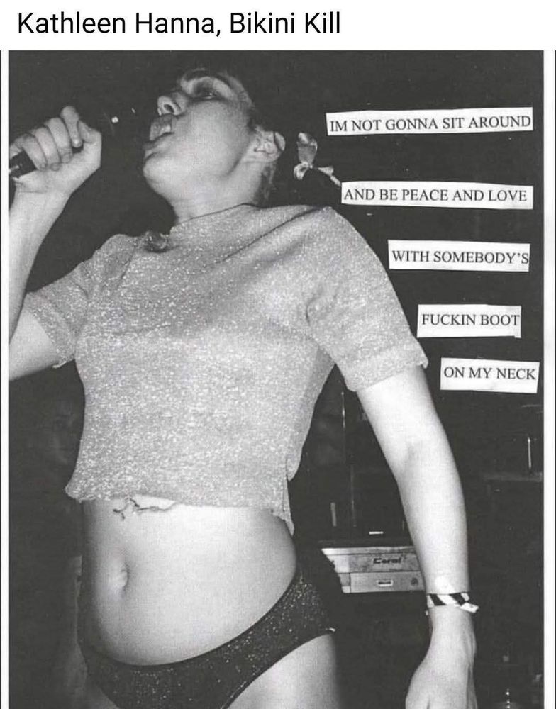 Black-and-white photo of punk icon Kathleen Hanna mid-performance with Bikini Kill, holding a microphone close to her mouth while passionately singing or shouting. She wears a glittery cropped top and black underwear-style bottoms, revealing a visible tattoo on her lower stomach. Her expression is intense, and her body language is defiant and energized. On the right side of the image, a collage-style cutout text reads: 