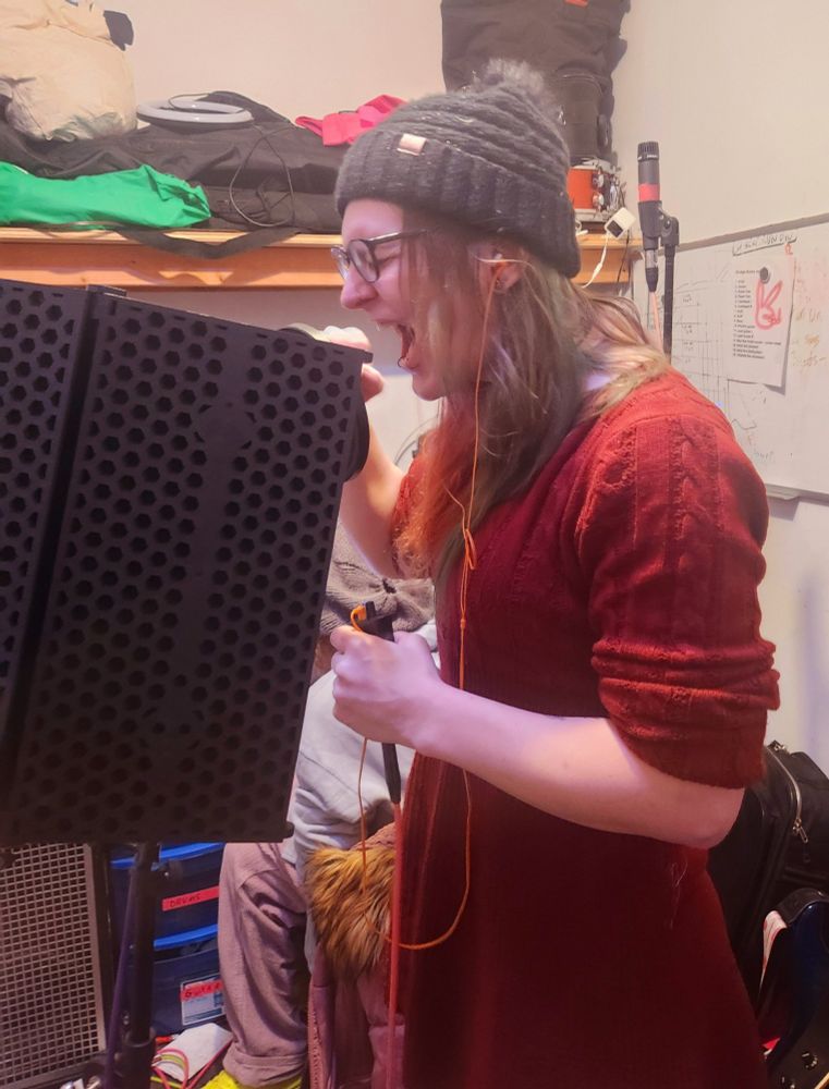 A woman in winter clothing screams into a microphone in a DIY studio setting.