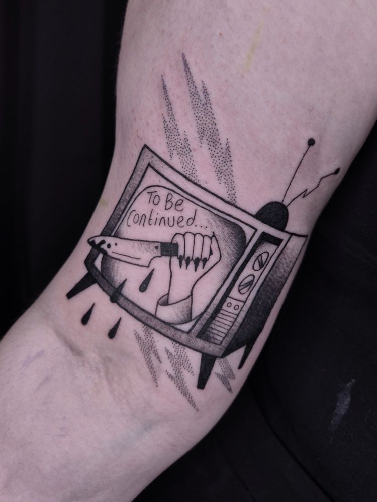 A black ink tattoo, on the inner bicep, of an old fashioned TV, on the screen there us a hand holding a knife, dripping with blood, and the words 'to be continued...' just above it 