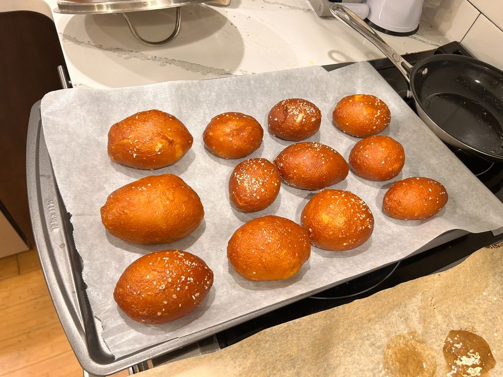 A baking tray, parchment-papered, full of somewhat misshapen pretzel rolls sprinkled with salt. Some lye stains are visible on some more parchment paper in the corner.