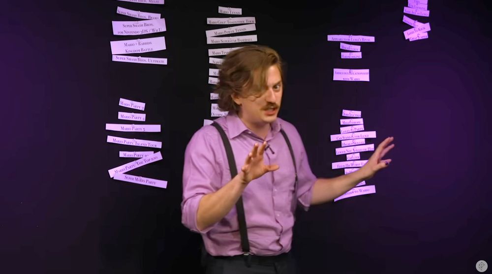 A man with brown hair wearing a light purple button up with black suspenders standing in front of a black wall with white papers on it lit with purple lighting. It is an image of Brian David Gilbert, from "Waluigi, Unravelled."