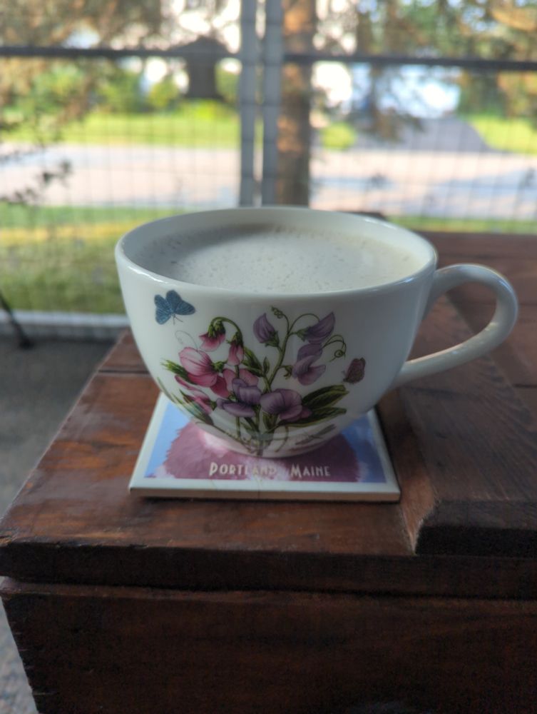 A large tea cup with sweet peas painted on it filled with earl grey, streamed oat milk, and a spoonful of maple syrup. It's sitting on a wooden trunk in the background you can see it's a lovely morning outside